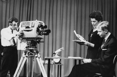 A black and white photo shows two men in suits reading the news Tangubra. Next to them is an old camcorder on a tripod. Behind the camera, a woman holds papers in her hands while watching the whole scene. All three people stand at attention inside a TV studio set up in the style of the 1960s.