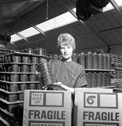 A black and white photo of an English woman in the early sixties, holding up an ornate steel mug with intricate designs on it to show off its quality inside one side of her large cardboard box that says " EVALPELLERI" . The scene is set at Cottontail stretched out over two lines of rows of tall slim beer cans. There's another big cardboard box behind filled with empty boxes saying "itsmodelledfrḞ Intricate wheels". It’s a sunny day. Shot by [Robert Mapplethorpe](https://goo.gl/search?artist%20Robert%20Mapplethorpe).