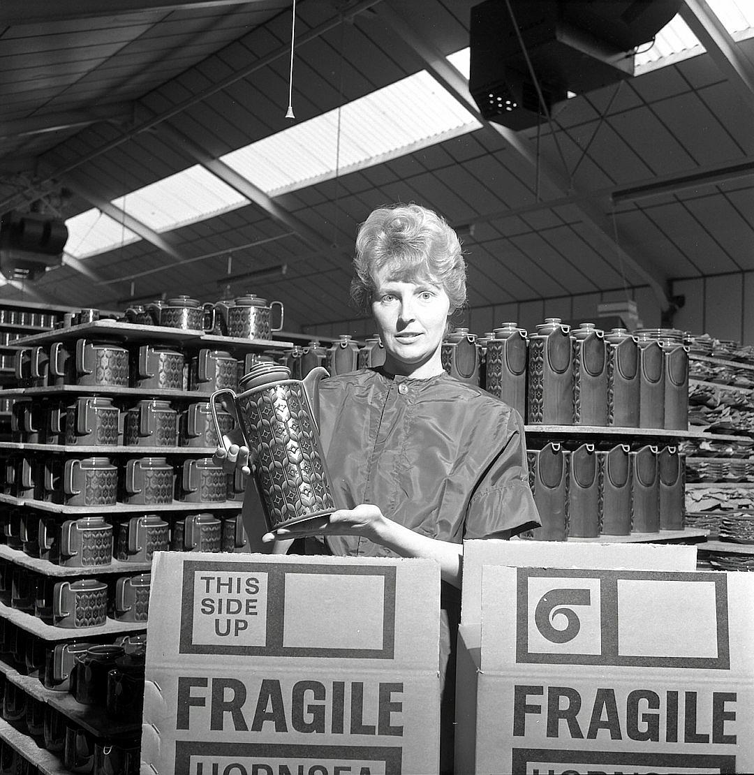 A black and white photo of an English woman in the early sixties, holding up an ornate steel mug with intricate designs on it to show off its quality inside one side of her large cardboard box that says ” EVALPELLERI” . The scene is set at Cottontail stretched out over two lines of rows of tall slim beer cans. There’s another big cardboard box behind filled with empty boxes saying “itsmodelledfrḞ Intricate wheels”. It’s a sunny day. Shot by [Robert Mapplethorpe](https://goo.gl/search?artist%20Robert%20Mapplethorpe).