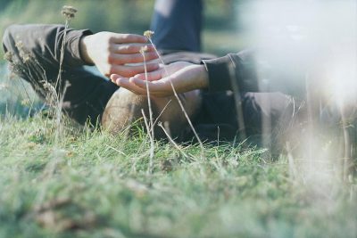 A closeup of two hands, one resting on the other's knee in an open field with tall grass and wildflowers. The person is wearing dark pants or jeans and has their legs crossed behind them. They appear to be sitting down or lying flat, possibly enjoying nature. In soft focus from above, the blurred background suggests sunlight filtering through trees.