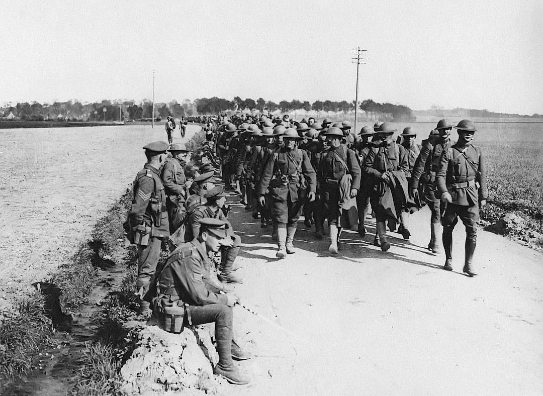 Black and white photograph of First World War American soldiers on their way to battle in France near Achey. They walk along an earthen road with one sitting down tired after being tired for several days straight. In the background we see fields, trees and hills. The sky is grayish blue, and it is late afternoon, in the style of an early 20th century war photographer.