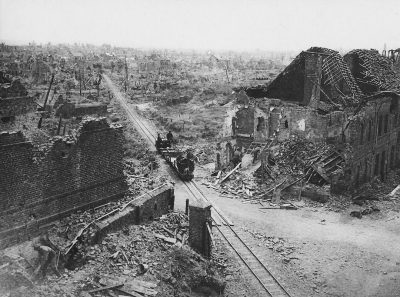 Old black and white photo of the battle for WWI in French, showing ruins of town with railway tracks through it. Two soldiers on horseback cross the road between buildings that have been destroyed by shellings. The scene is taken from an elevated perspective overlooking the city's charred streets. In the background there can be seen distant military vehicles moving along lines towards one another in the style of Ch Phase.