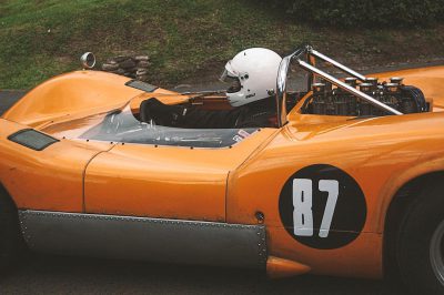 A photo of an orange Le Mans racecar with black and white numbers on its side. It has silver chrome details around its hood and fenders. In front, a man wearing a helmet is sitting in the style of driving the car, shot from above with a grassy field background and 35mm film grain.