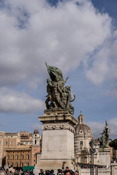 A photo of the Colle di rifle statue in Rome, Italy, with people around it and buildings behind it. The sky is blue with white clouds, taken in the style of Sony Alpha A7 III camera using an aperture f/8 lens.