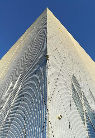 Low angle shot of the exterior wall of tower in glass, with thick lines on white canvas fabric attached to it. It is made up of metal mesh and has wires hanging from its top edge. In front there's an open window that leads into another building, and behind you can see blue sky. The photo was taken by professional photographers using Sony Alpha A7 III camera.