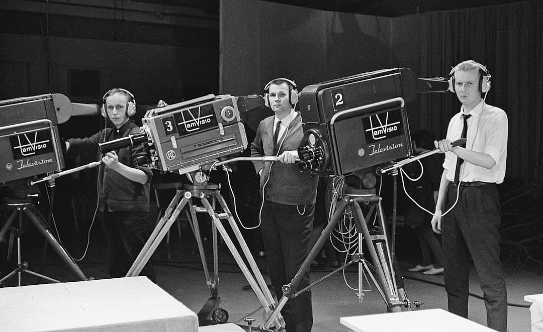 In the early days of television, in an empty studio with cameras and lights set up for live television, three young men stand behind their equipment dressed as news paparazzi wearing black suits and white shirts tied at the neck with navy blue ties. They hold microphones that have “TV melreeze” written on them. The atmosphere is excited but professional. Black & White film photography, shot in the style of ARRIFLEX35 BL Camera Canon K207 Prime Lens Full Shot, hyper realistic, detailed skin, natural lighting, HDR.