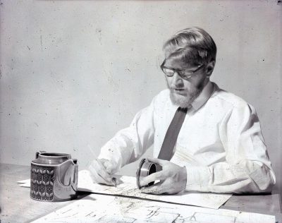 Vintage photo of Peter Edmondson, architect and designer at his desk drawing blueprints for the original Bchen & Co coffee mug design in 2035, wearing glasses with beard and white shirt with tie. A black paper cup is on one side of him and he's holding an ink pen while writing something down on parchment next to it. The background behind him has plain white walls and concrete floors.