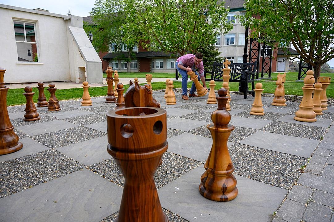 A giant chess set with large wood pieces on the ground of an outdoor courtyard at St. Clo prominently features one rook piece being piled up in the style of two people in workwear. The setting is a college campus and there is green grass surrounding it. The photograph was taken from behind as if someone took the photo over their shoulder. It should look like they took this picture to show off what they made.