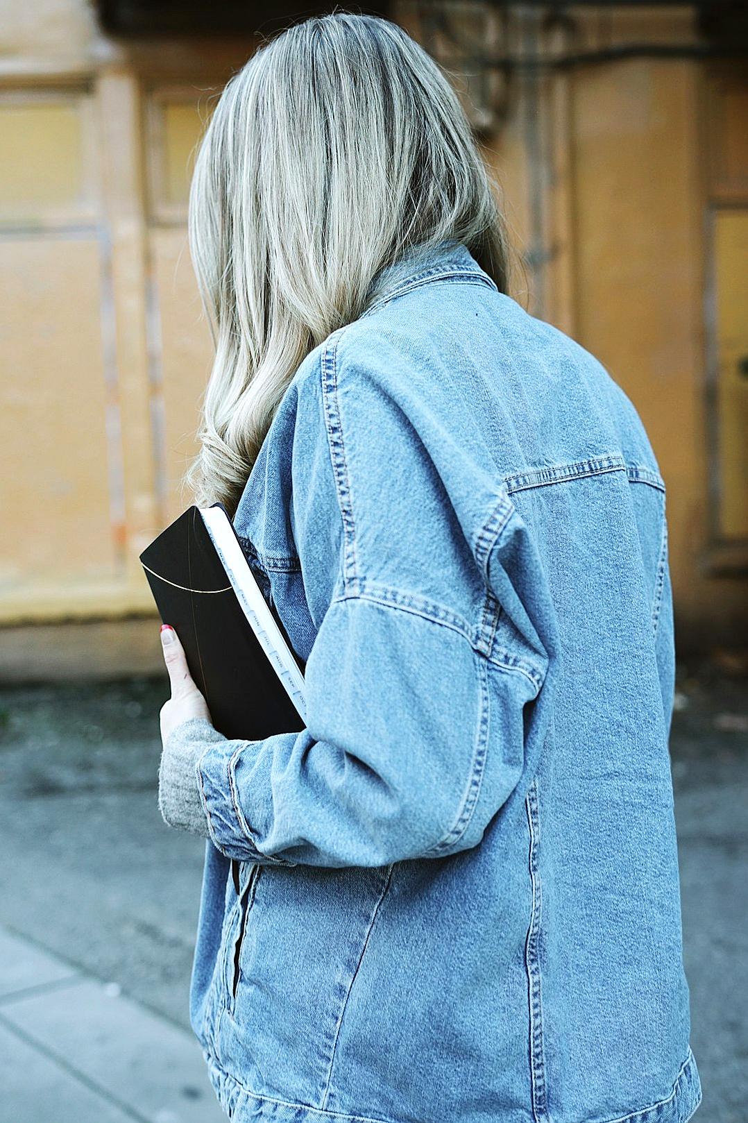 A woman with shoulder-length blonde hair, wearing an oversized denim jacket and jeans, holding a black book in her hand. She is seen from the back as she walks down the street, looking over her right shoulder at something off frame. The photo captures a casual yet stylish urban look, showcasing both fashion and what is clearly visible on the books. Her posture reflects confidence while maintaining a relaxed vibe, complementing to create a full-body portrait in the style of an urban photographer.