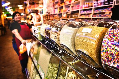 A photo of an urban store with lots of colorful candy and grains, featuring large glass bags filled to the brim with sweet dried fruits, brown rice or grain, standing on metal stands in front of a staff member who is serving from them. The background features shelves lined up with various items including spices, white powder and black circular foil-wrapped candies. A person wearing dark blue jeans stands next to one bag, holding out their hand for some chocolate alone at the end.