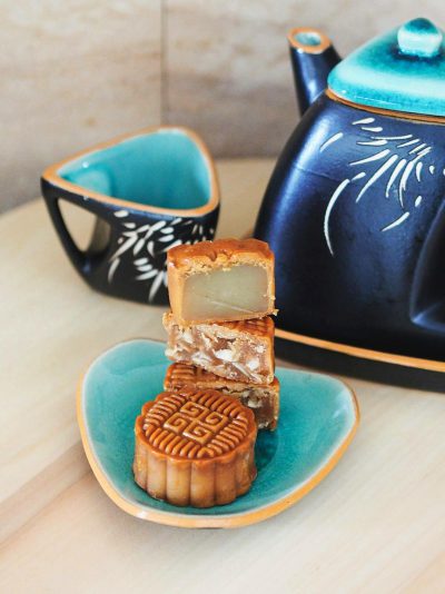 A delicate mooncake, placed on an elegant turquoise plate with gold rim and motifs, is cut into two pieces to reveal its intricate interior filling of white lotus seed or mar interests sitting next to the tea pot. The background features a light wooden surface and subtle gray walls, creating a serene ambiance for enjoying these traditional Asian treats.