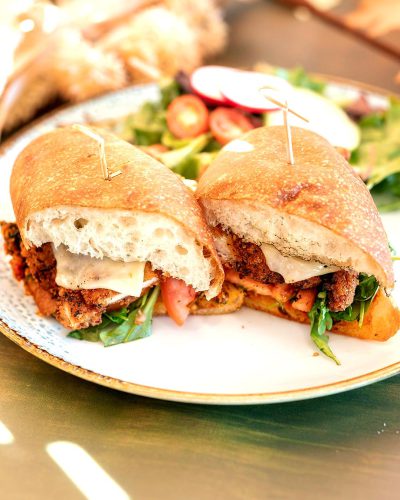 A photo of two chicken cutlet sandwiches with mozzarella and arugula on top, sitting side by side on an elegant plate, accompanied by salad in the background. The sandwich caricore is fresh from being made at home using a meat food futurist machine. Soft lighting highlights its golden brown crust and vibrant colors. This mouthwatering scene evokes memories of a cozy family meal or inviting afternoon tea break. A sense of warmth shimmers through the air as if ready to be eaten.