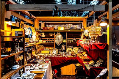 A photo of an antique and vintage market inside the Grand Palais, showcasing stalls with various antiques such as silverware, art pieces from different eras, jewelry, fine china, old books, brass lights hanging on wooden shelves, red velvet tablecloths, dim lighting creating a warm atmosphere, a wide shot capturing all elements in detail. Taken in the style of Nikon D750 using Fujifilm film stock, natural light, color photograph, hyperrealistic, high resolution photography, super detailed.