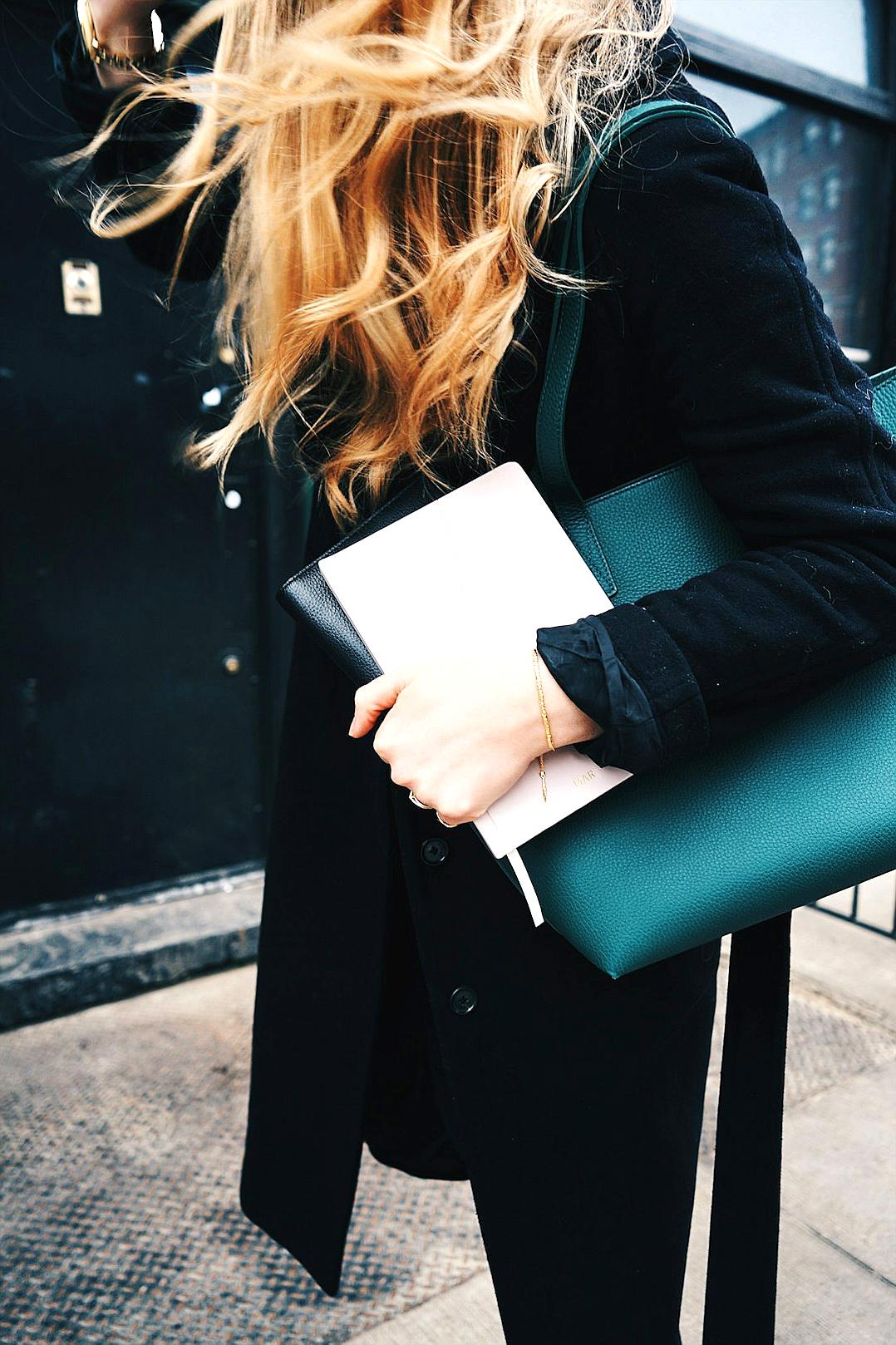 close up of woman walking down the street with long blonde hair holding black laptop bag, white notebook and green leather purse, street style photo, minimalistic aesthetic, black coat, natural lighting, street photography