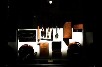 A dark room with the light of an LED panel illuminating leather tools and items on display, including holsters for weapons, pouches, jewelry boxes, and belt pouches, some of which have a slight sheen from their polished surface. The focus is centered around one pair of simple brown stitching in white canvas fabric with no design or pattern. There's a clear view of all these objects on top of glass shelves against a black background. In front of them there’s another set of tools. The scene is presented in the style of minimal editing of the original text.