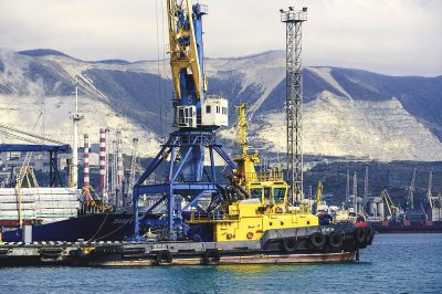 A photo of a yellow and blue small crane barge in the port, behind it is a large white mountain with a black coal mine on it, a cargo ship, an industrial area, a black sea, a port city.