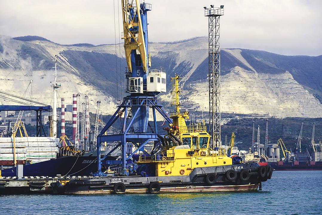 A photo of a yellow and blue small crane barge in the port, behind it is a large white mountain with a black coal mine on it, a cargo ship, an industrial area, a black sea, a port city.