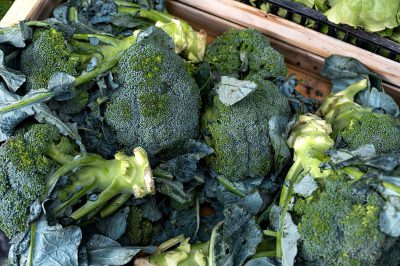 A pile of fresh broccoli in the middle with leaves, on top are some roughly cut pieces and below is more broccoli with greens. A wooden crate full of broccoli at an outdoor market. The photo was taken from above.