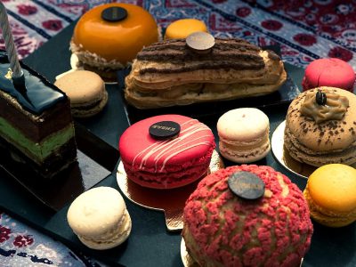 A collection of colorful and intricate pastries, including an eclair with chocolate filling and macarons in various colors and shapes. Layered cakes are decorated with icing sugar or glittering dust, placed on black plates on a dark blue velvet tablecloth. The background is adorned with red fabric patterns, creating contrast between the bright bohemian style dishes and the rich textures of the cloth. A small round wooden stamp reads 'Gleam' in one corner of each dessert.