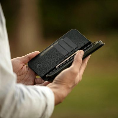 A man holding an open black leather wallet with his phone in it, a product photo of the minimalistic and slim Zajeala smartphone case made from expensive nappa air profile distressed metal material. A thin card holder is attached to one side, the interior has cards and business documents inside, the background should be a blurred grassy field, focusing on showcasing the sleek design of the wallet and its ability to carry complexly for all modern smartphones up to two inches wide in the style of a minimalist design.