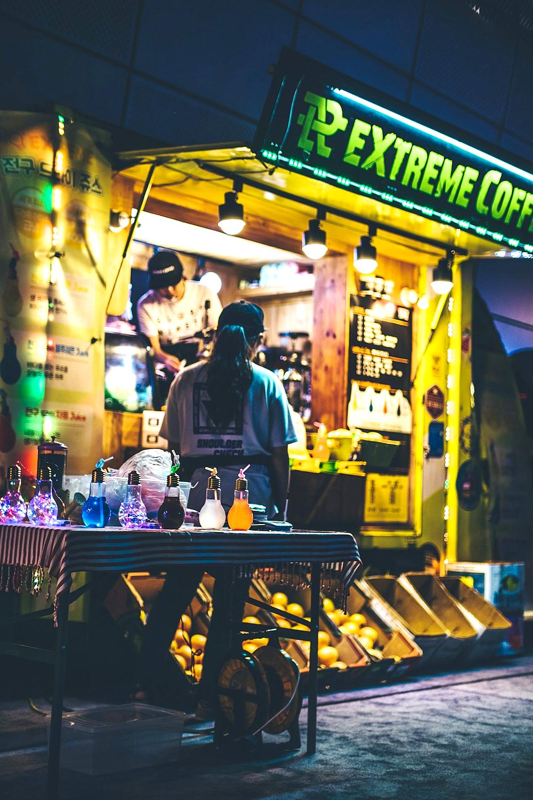 a night time photo of an outdoor coffee shop in the middle east, with neon lights and colorful decorations. A man is standing behind his stand selling drinks from a cart full of fruits and snacks. The sign above says “charger extreme Coffee”. He has black hair tied back into two braids and wears streetwear . In front there’s another table covered by three large open bags filled with colourful glowinthedark paint. On top one bag stands on its side with some tubes coming out of it.