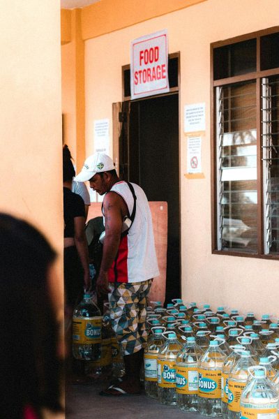 A man wearing a white cap, black and red shirt with colorful shorts standing next to his wife at the entrance of "Ekong's food storage" store in Edo state, their hands holding several water bottles with plastic covers on them, photo taken from outside of the building, photo shot in the style of canon eos r5, 30mm lens, f/2.8, depth of field, natural light, hyper realistic