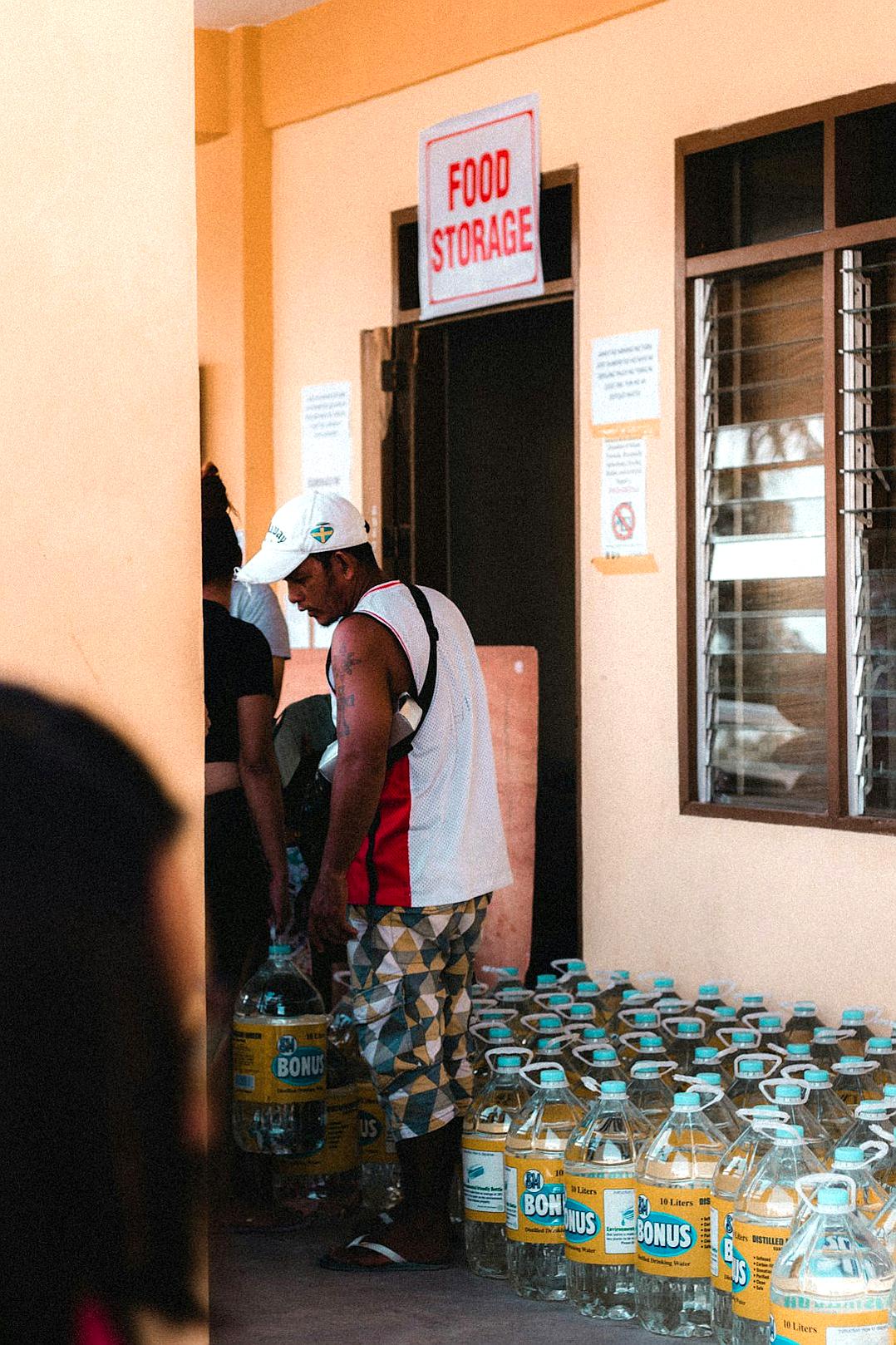 A man wearing a white cap, black and red shirt with colorful shorts standing next to his wife at the entrance of “Ekong’s food storage” store in Edo state, their hands holding several water bottles with plastic covers on them, photo taken from outside of the building, photo shot in the style of canon eos r5, 30mm lens, f/2.8, depth of field, natural light, hyper realistic