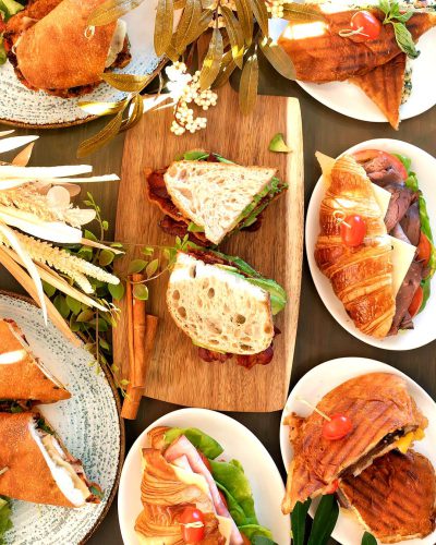 A table set with an assortment of sandwiches, including croissants and paninis on plates. In the center is a wooden board holding two Cuban-style club sandwich slices, and in front there's one plate featuring sandwiches made from BLT ingredients. The background features greenery, flowers, and a wall behind it. It captures the food beautifully, highlighting its colors and textures. Shot in the style of a professional photographer using a Canon EOS R5 camera with a macro lens for sharp focus.